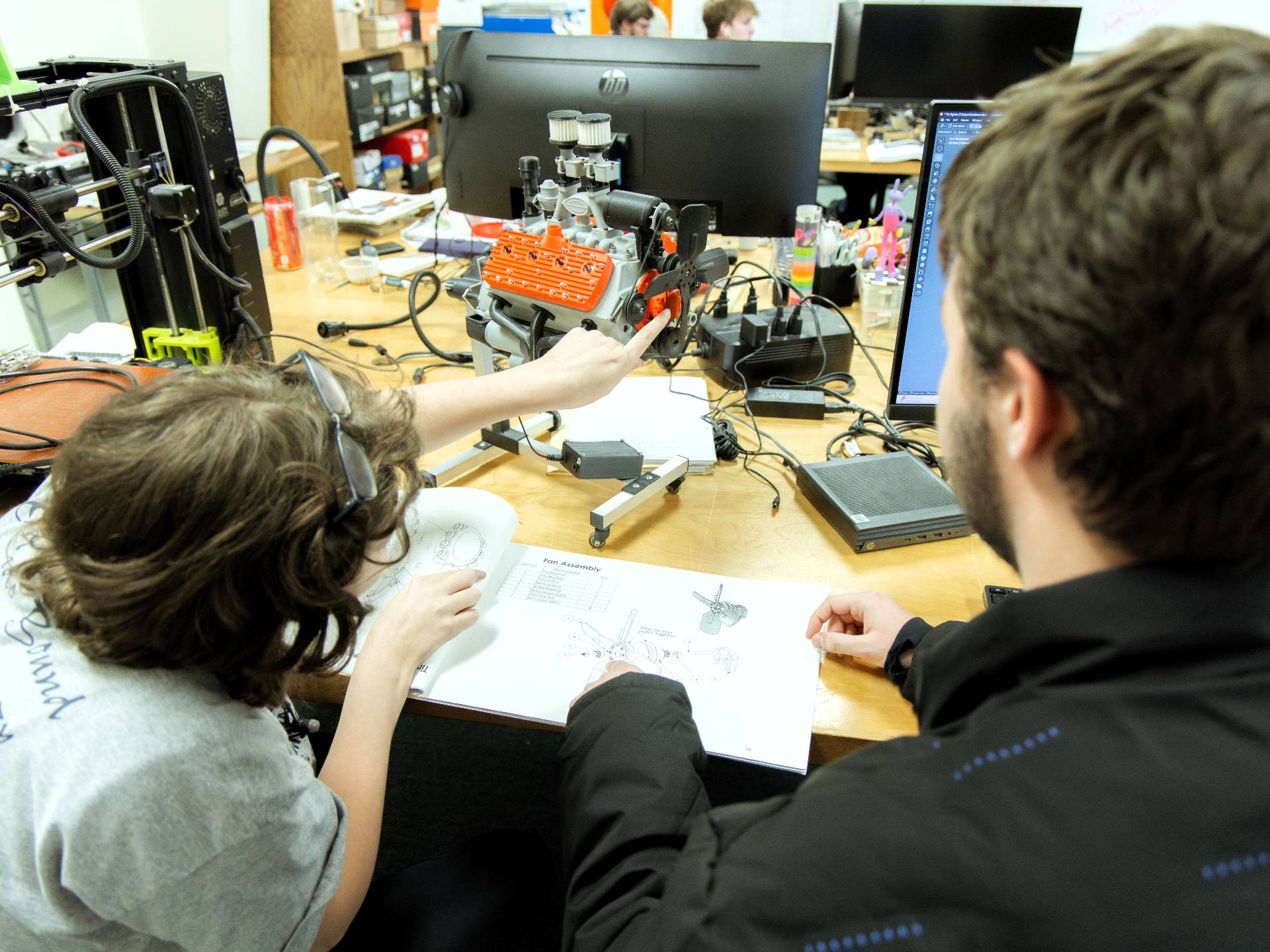 A man and a younger person sit at a desk in an office, looking at an instruction manual while working on a small model engine. Computers and equipment are visible in the background