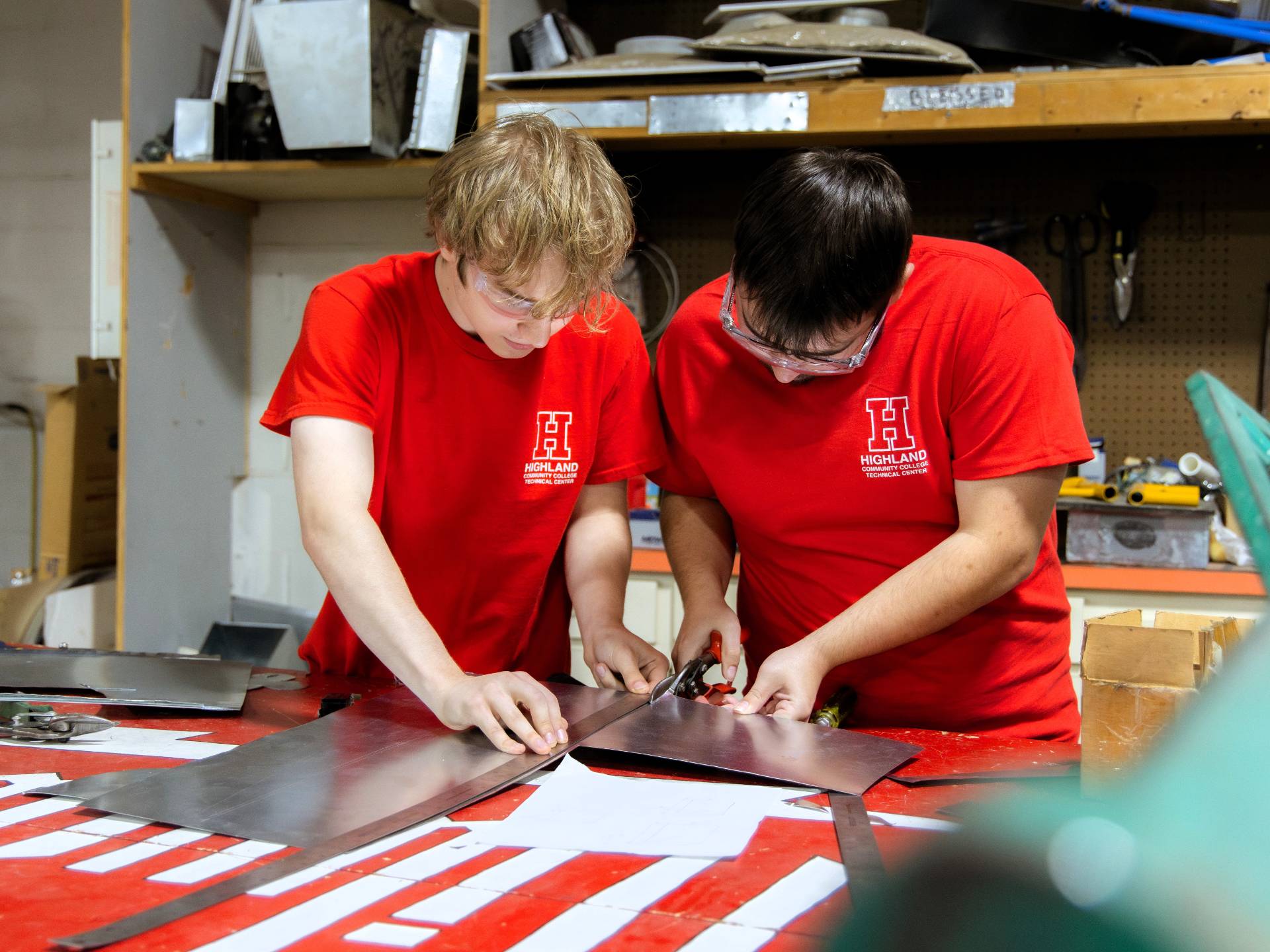 Two young men wearing red Highland Technical College t-shirts and safety glasses work together to cut a piece of sheet metal with tin snips in a workshop setting.