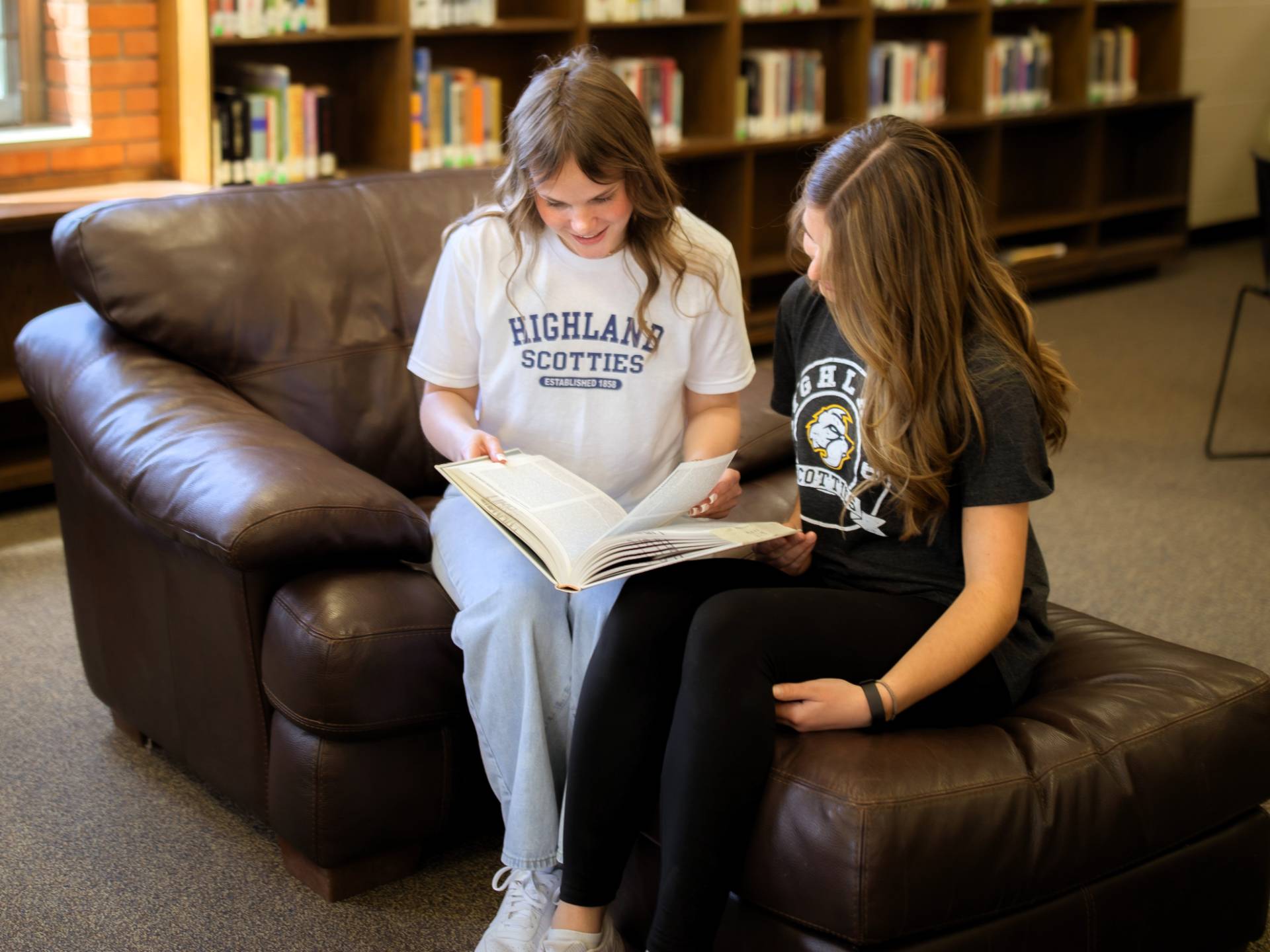 Two students are sitting on a coach in a library reading a book together. 