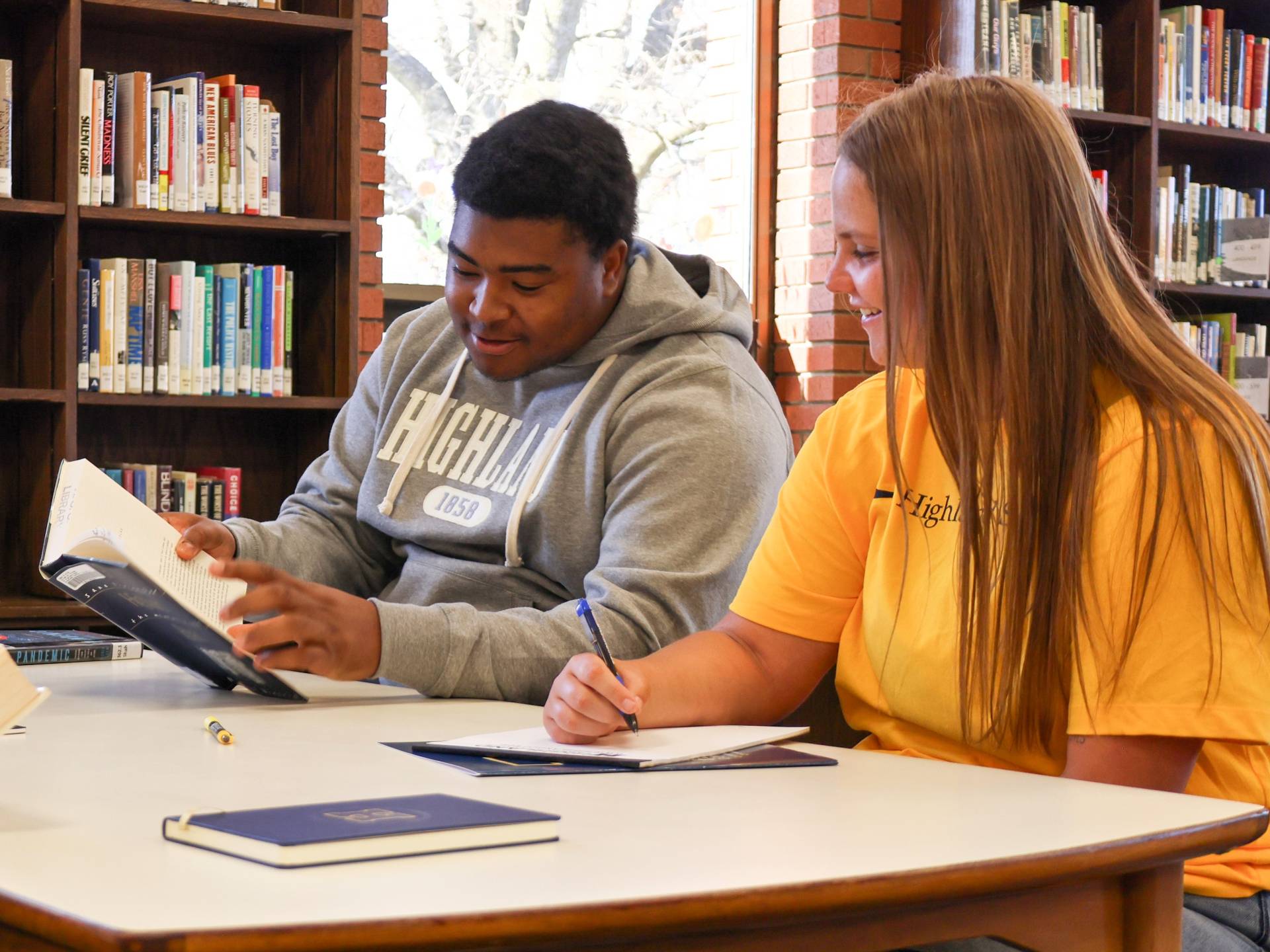 Two students are studying in the library, looking at a book together. 
