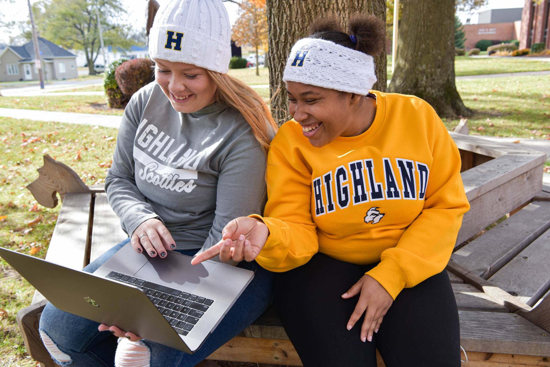 Two students sitting outside under a tree, one student is holding a laptop while the other is pointing at the screen